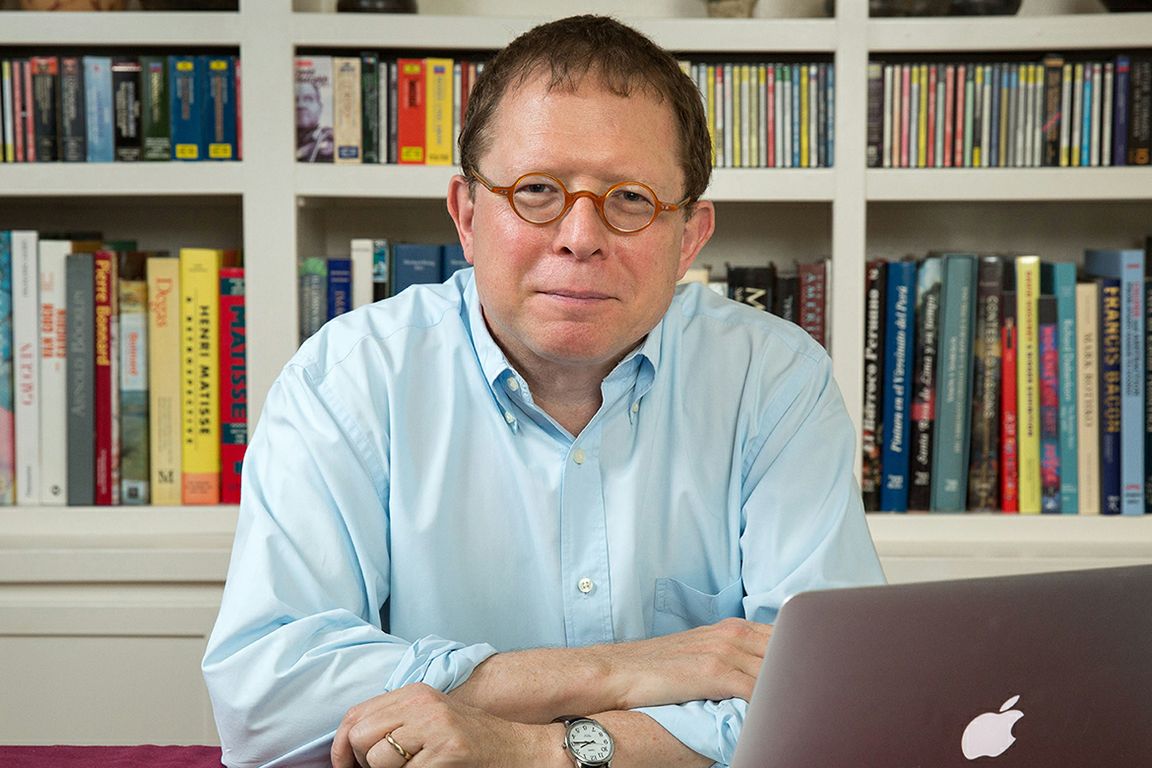 Efrain Kristal sits at a desk with a laptop, wearing a light blue shirt and round tortoiseshell glasses, with a large bookshelf behind him. 