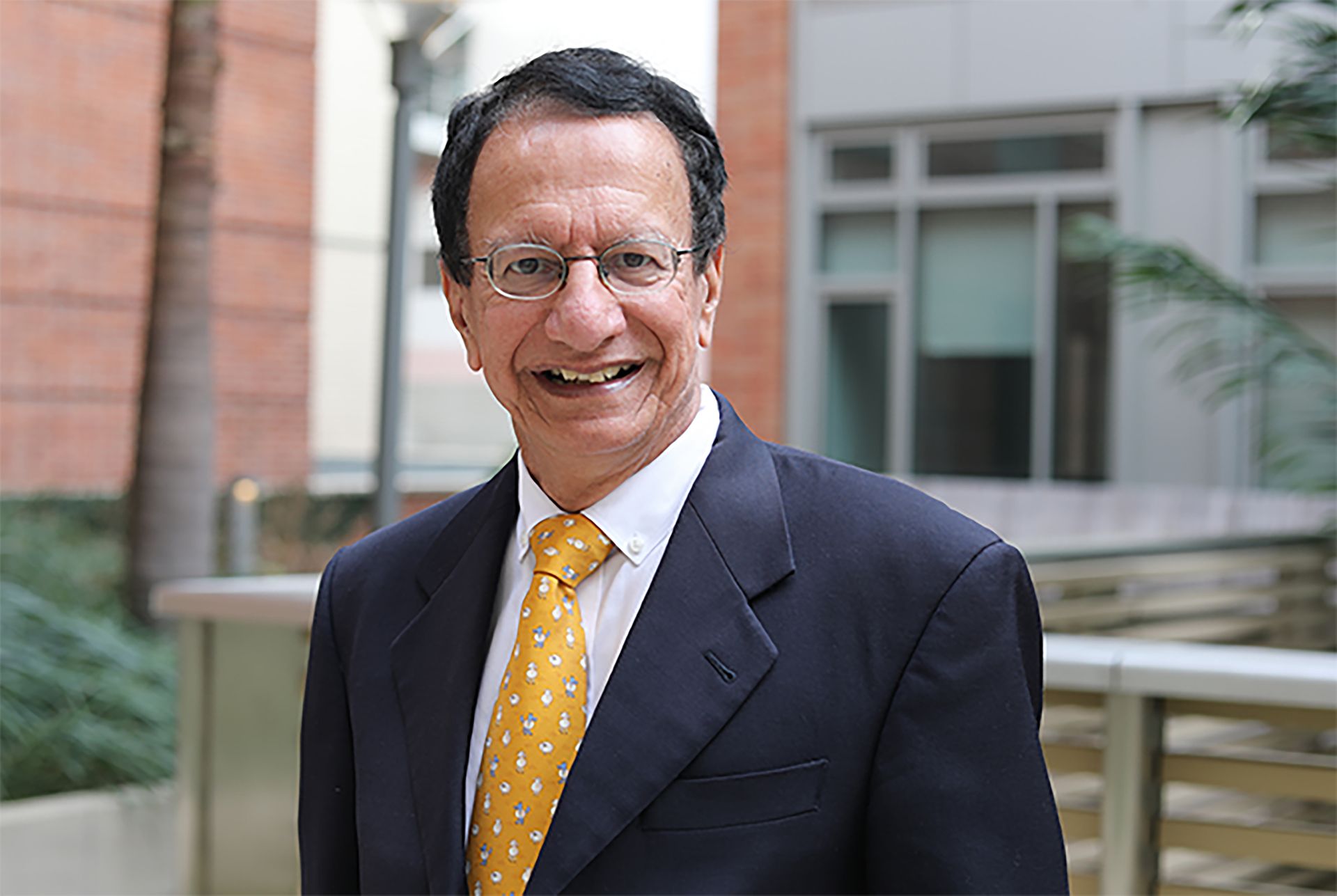 Headshot of Chandrashekhar Joshi smiling, wearing a dark suit and gold tie, with a modern building in the background.