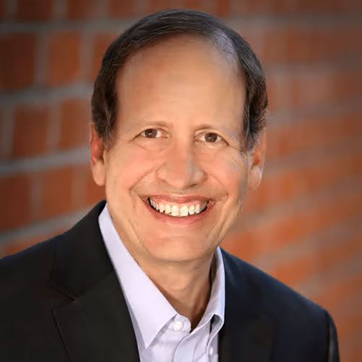 Headshot of Michael Galper smiling, wearing a black blazer and light purple dress shirt, with a brick wall in the background.