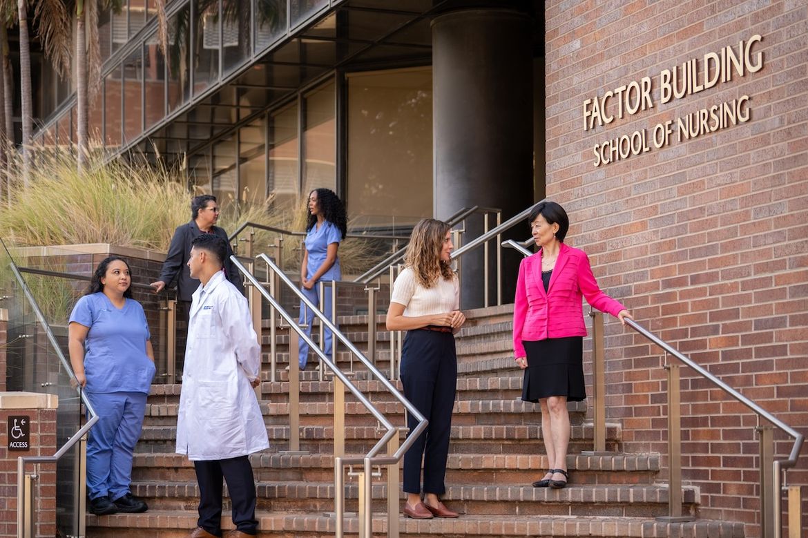 Several students and faculty members in scrubs and professional attire gather on the exterior steps of the Factor Building, School of Nursing at UCLA.