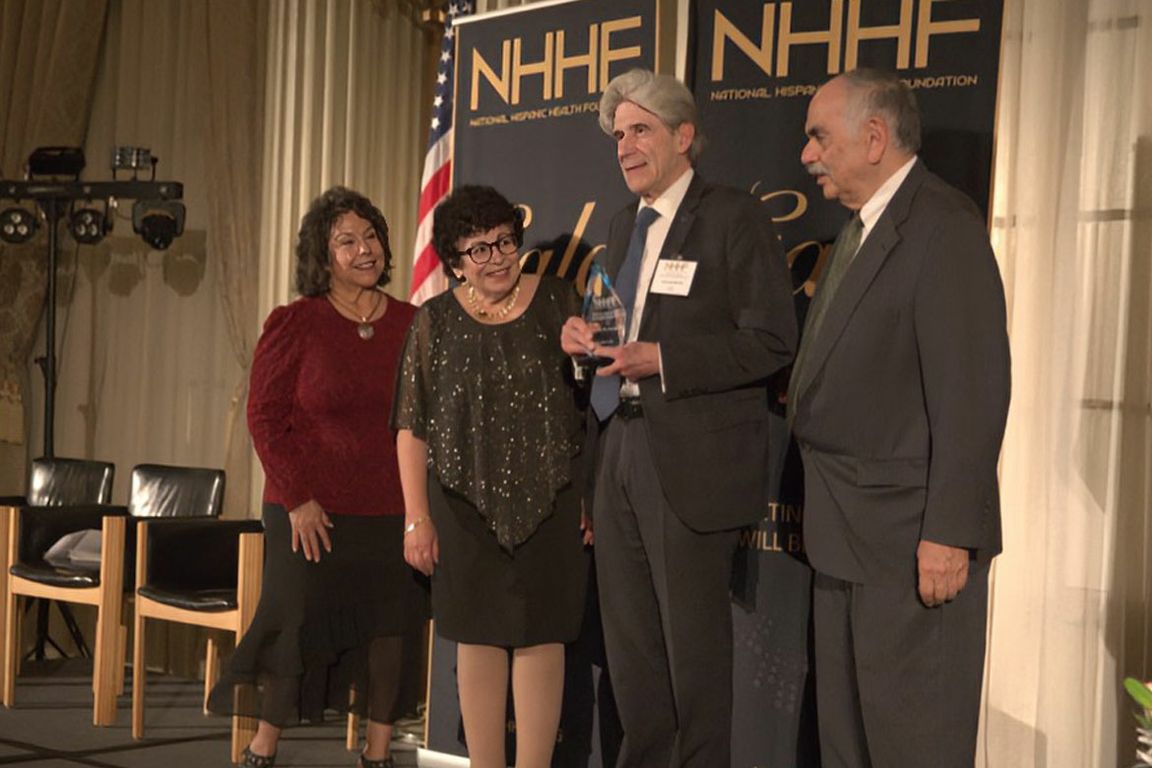 UCLA Chancellor Julio Frenk accepts a glass award trophy on stage at a National Hispanic Health Foundation (NHHF) event, flanked by three colleagues in formal attire.