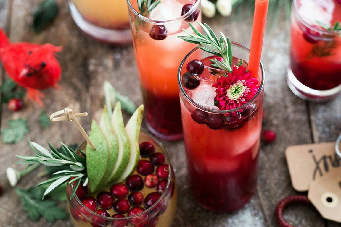 An overhead view of festive drinks garnished with cranberries, pear slices, rosemary sprigs, and a pink flower, arranged on a rustic wooden surface.