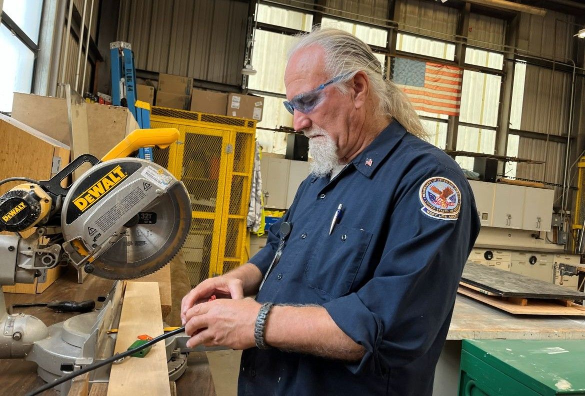 Daniel Martin, wearing safety glasses and a navy uniform, works at a woodworking bench beside a DeWalt miter saw in a workshop at the VA West Los Angeles campus, with an American flag visible in the background.