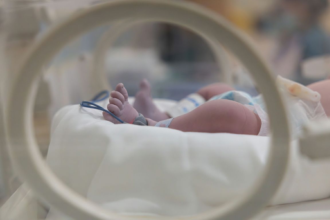 A premature newborn with a medical monitor attached to its foot rests in a hospital incubator.