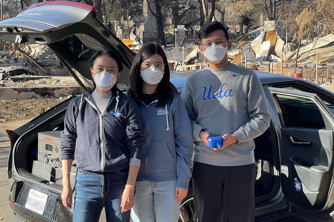Three researchers wearing N95 respirator masks stand together in front of a car with its trunk open, containing research equipment. Behind them is a neighborhood destroyed by fire, with burned debris and charred trees visible. Left to right: Yifang Zhu, Yuan Yao, and Haoxuan Chen