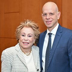 Joyce Brandman, wearing a light-colored tweed jacket, and Dr. John Belperio in a dark navy, suit stand together indoors, smiling at the camera in front of a wood-paneled wall.