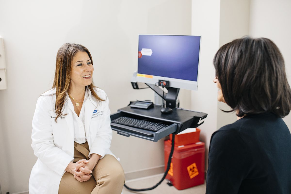 Dr. Sanaz Memarzadeh, wearing a white lab coat, smiles while speaking with a patient in a clinical exam room. A computer workstation and biohazard waste container are visible in the background. 