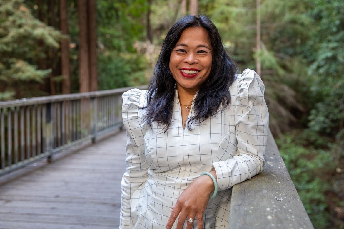 Celine Parreñas Shimizu in puffy white shirt stands with left arm on rail of wooden bridge amid redwood trees