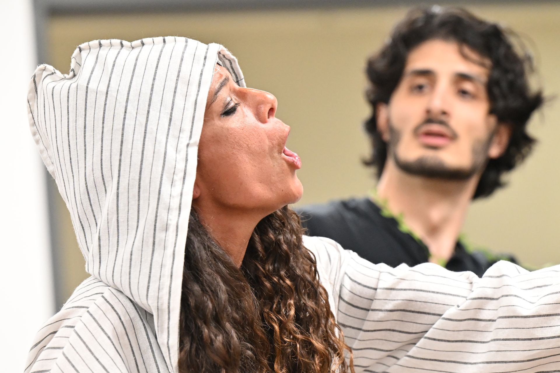 Woman in white-and-black striped hooded garment sings while another person stands in the background