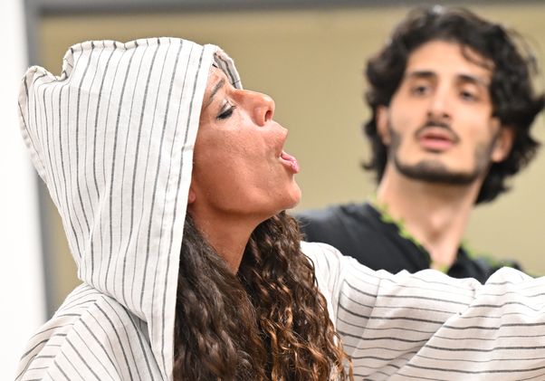 Woman in white-and-black striped hooded garment sings while another person stands in the background