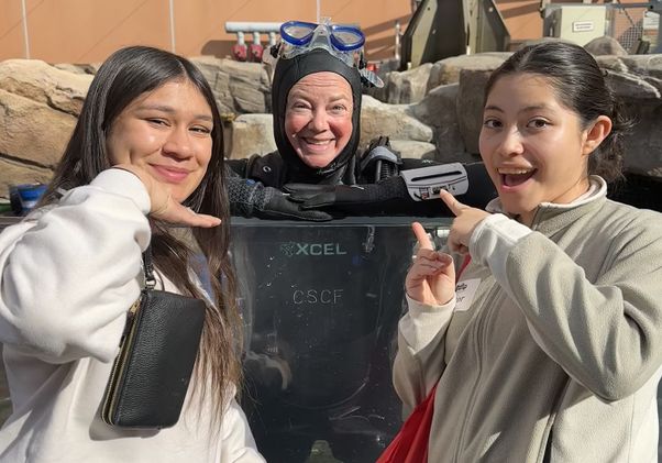 Two girls stand on either side of person in hooded wetsuit and diving googles
