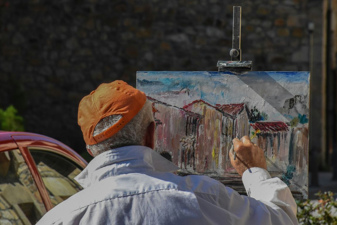 Older man in red hat paints a rustic building scene on a canvas easel outdoors, viewed from behind.