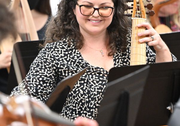Marylin Winkle, in black patterned dress and glasses, smiles as she plays a viola da gamba