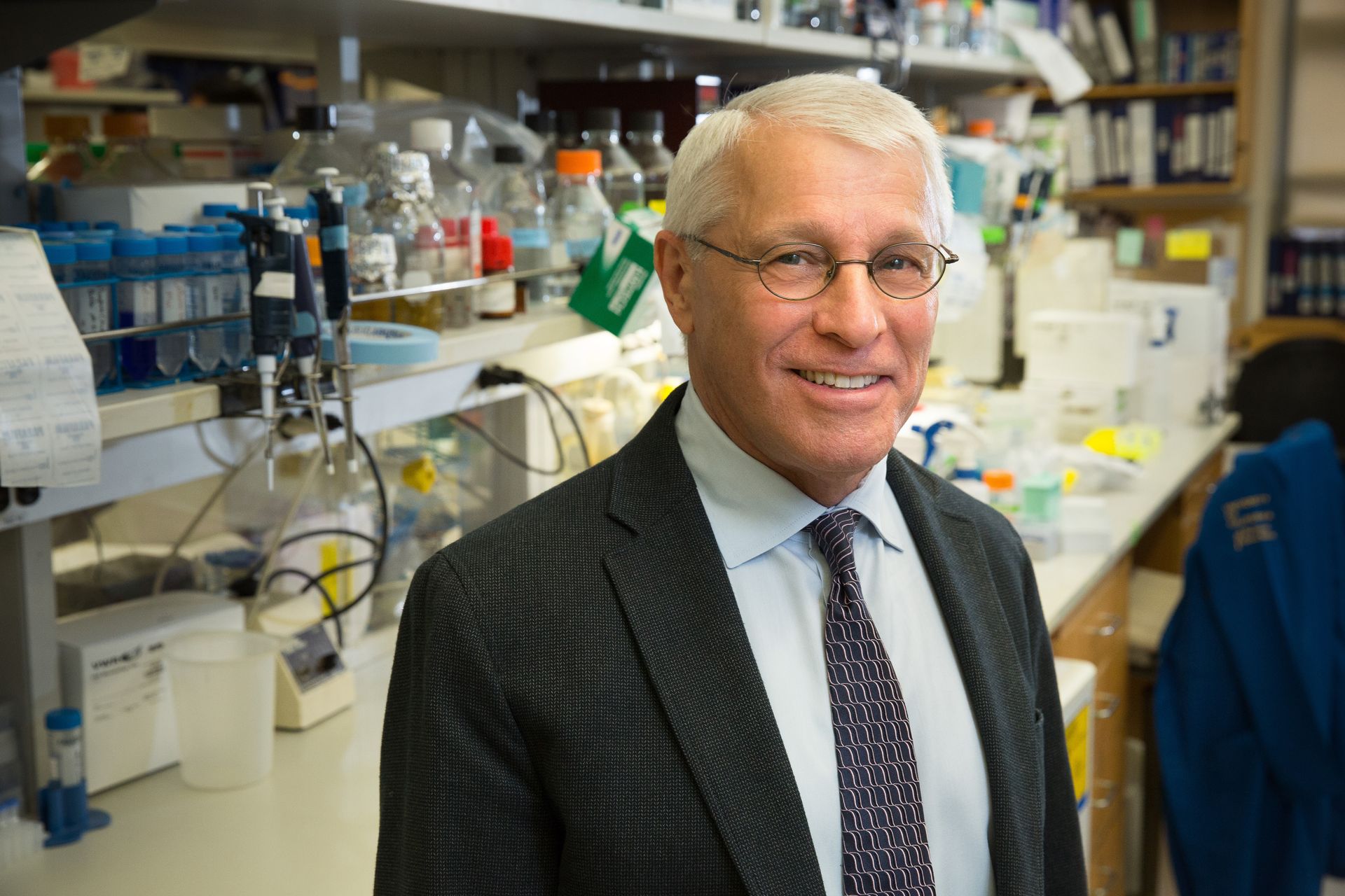 Owen Witte in glasses, suit and tie standing in front of lab equipment