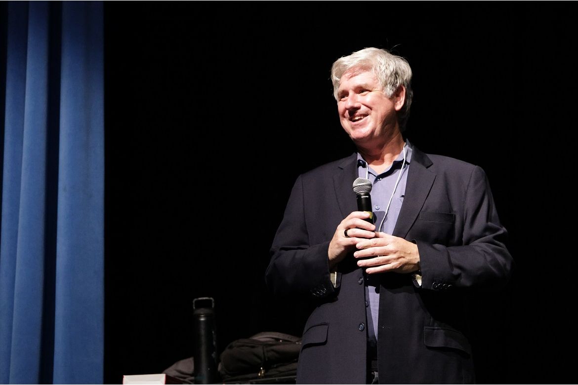 Robert Gould, in dark jacket and blue shirt, holding a microphone