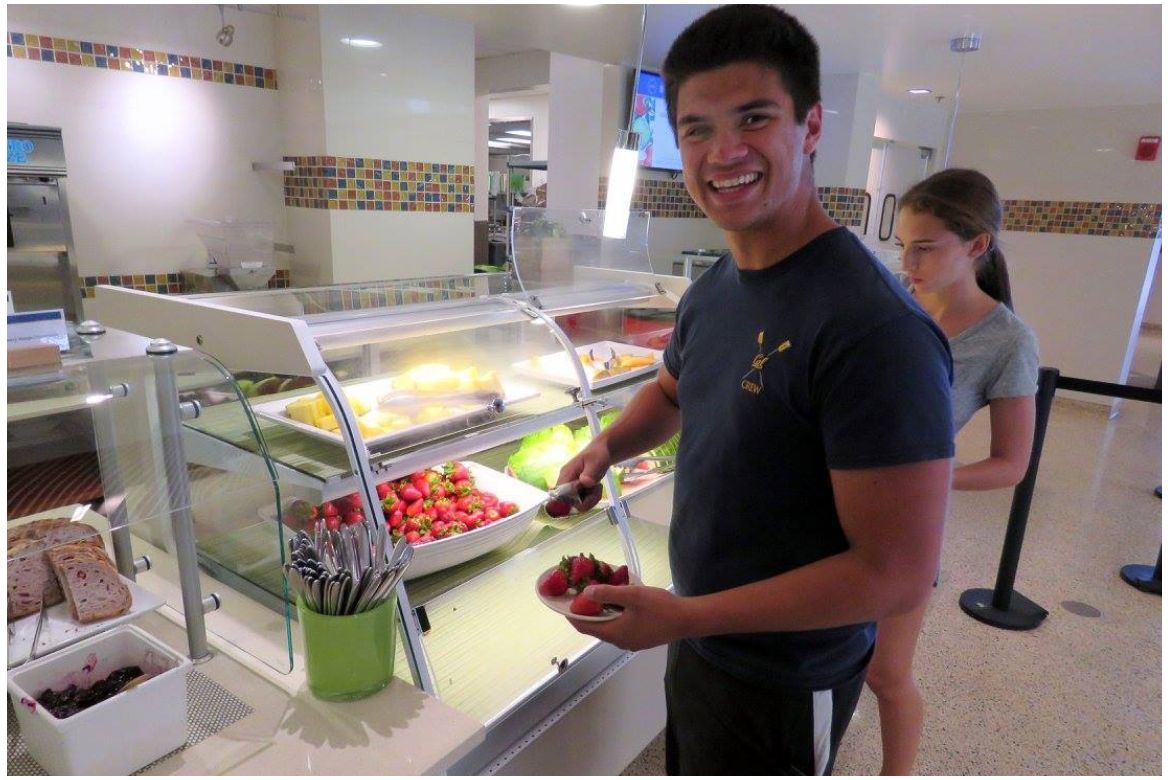 A male UCLA student in a buffet line holds a plate of four strawberries as he reaches for a big white dish full of more strawberries