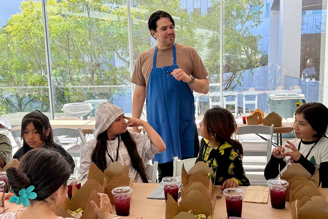 Chef Luis Sierra, standing in center in blue apron, speaks to students seated at a table