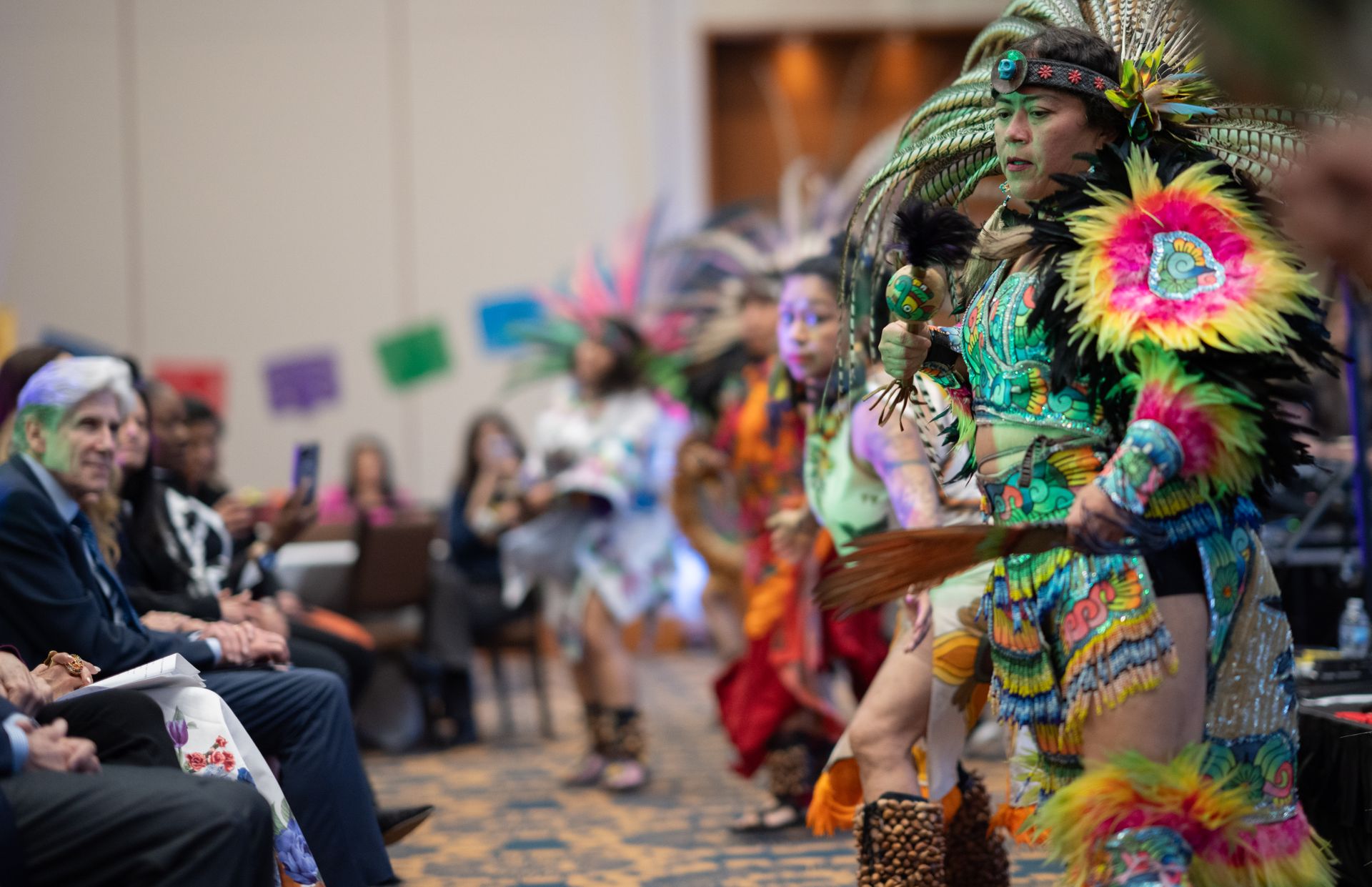 Aztec dancers in vibrant feathered regalia and colorful costumes perform at an indoor event before a seated audience, with papel picado decorations visible in the background.