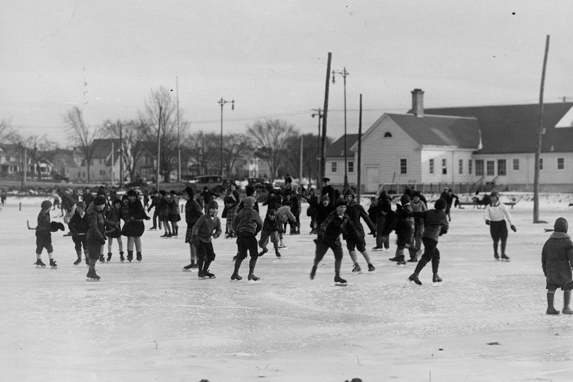 A black-and-white historical photograph of children ice skating on an outdoor rink in a residential neighborhood, with bare trees and a white building in the background.
