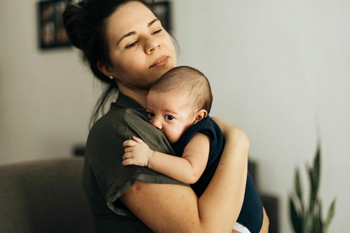 A woman with dark hair holds a wide-eyed infant against her shoulder in a home setting.