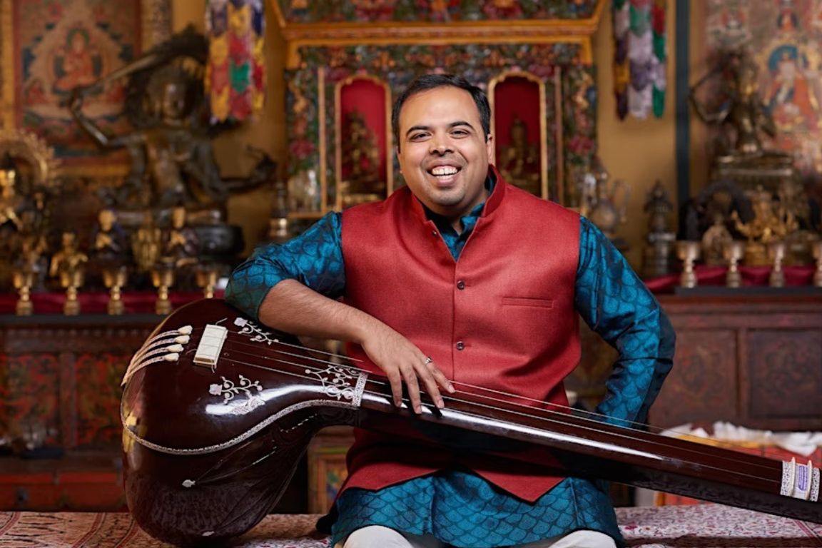 Samarth Nagarkar sits with instrument against colorful alter of Indian deities 