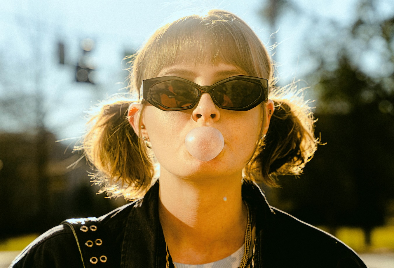 Bubble gum Close-up of a young person wearing narrow black sunglasses and pigtails, blowing a bubblegum bubble outdoors in bright sunlight, with a softly blurred street scene in the background.