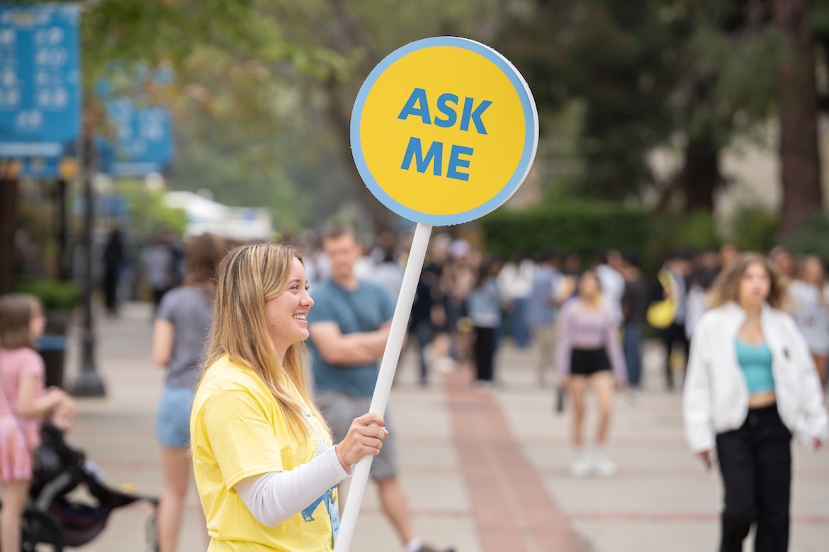 Bruin Day–Female volunteer in yellow UCLA shirt on campus, holding sign reading, "Ask Me."