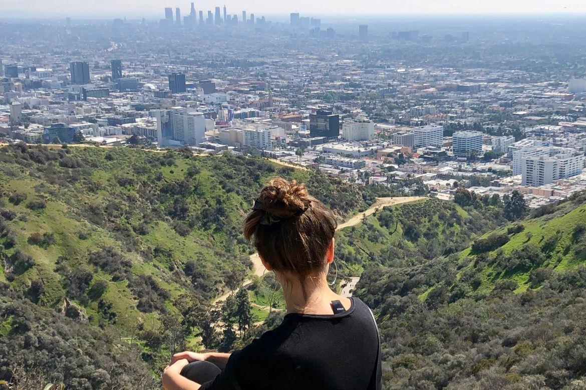 Female hiker seated on a rock atop a hill that overlooks the hazy Los Angeles city and skyline in the distance
