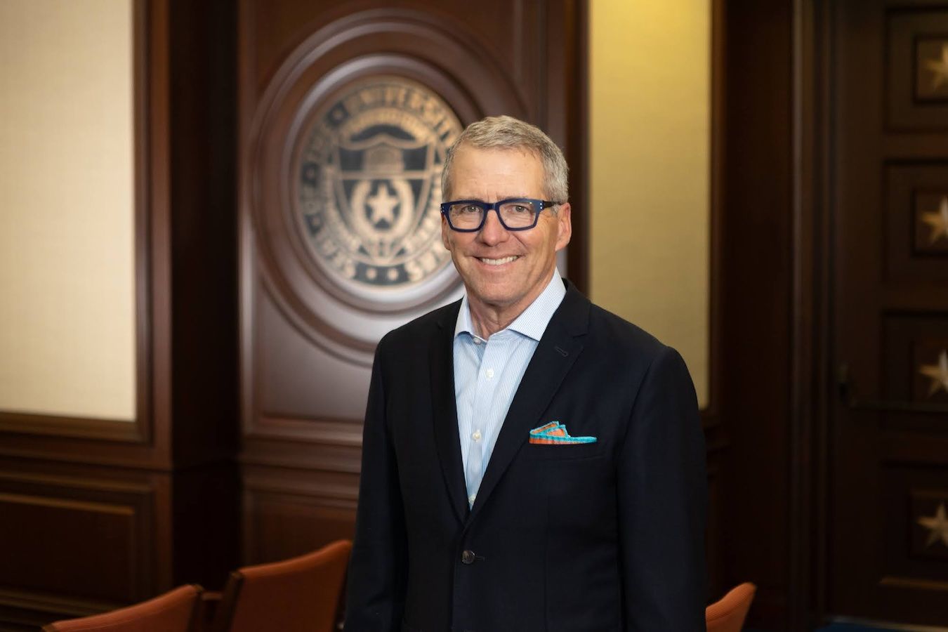 James B. Milliken, wearing blue-framed glasses, a dark blazer, and a light blue dress shirt with a colorful pocket square, smiles while standing in front of a wood-paneled wall bearing an institutional seal.