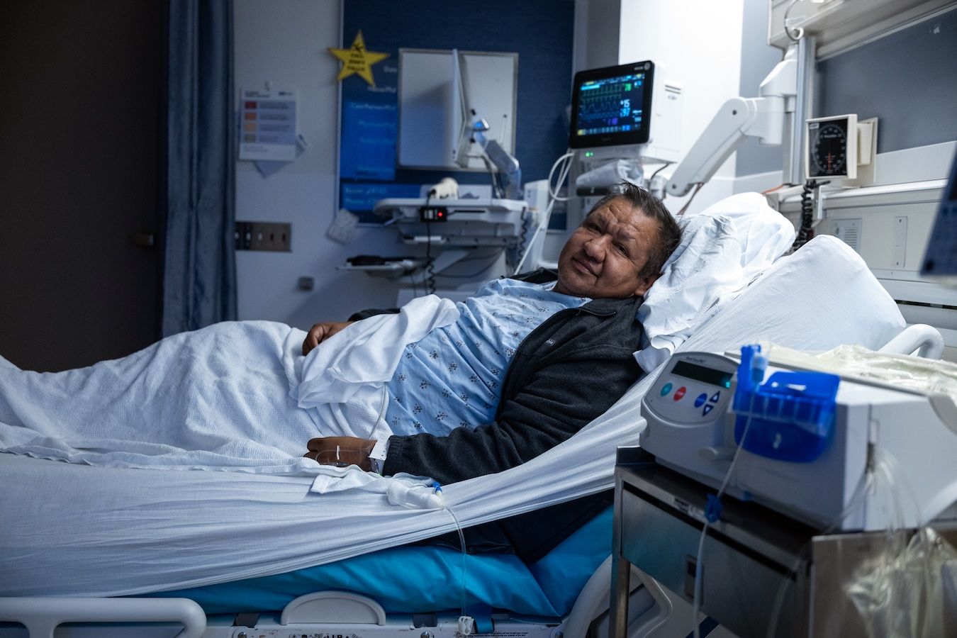 Oscar Larrainzar, the transplant recipient, rests in a hospital bed at UCLA wearing a patient gown and a dark jacket, with medical monitors and equipment visible in the background. He looks toward the camera with a calm expression.