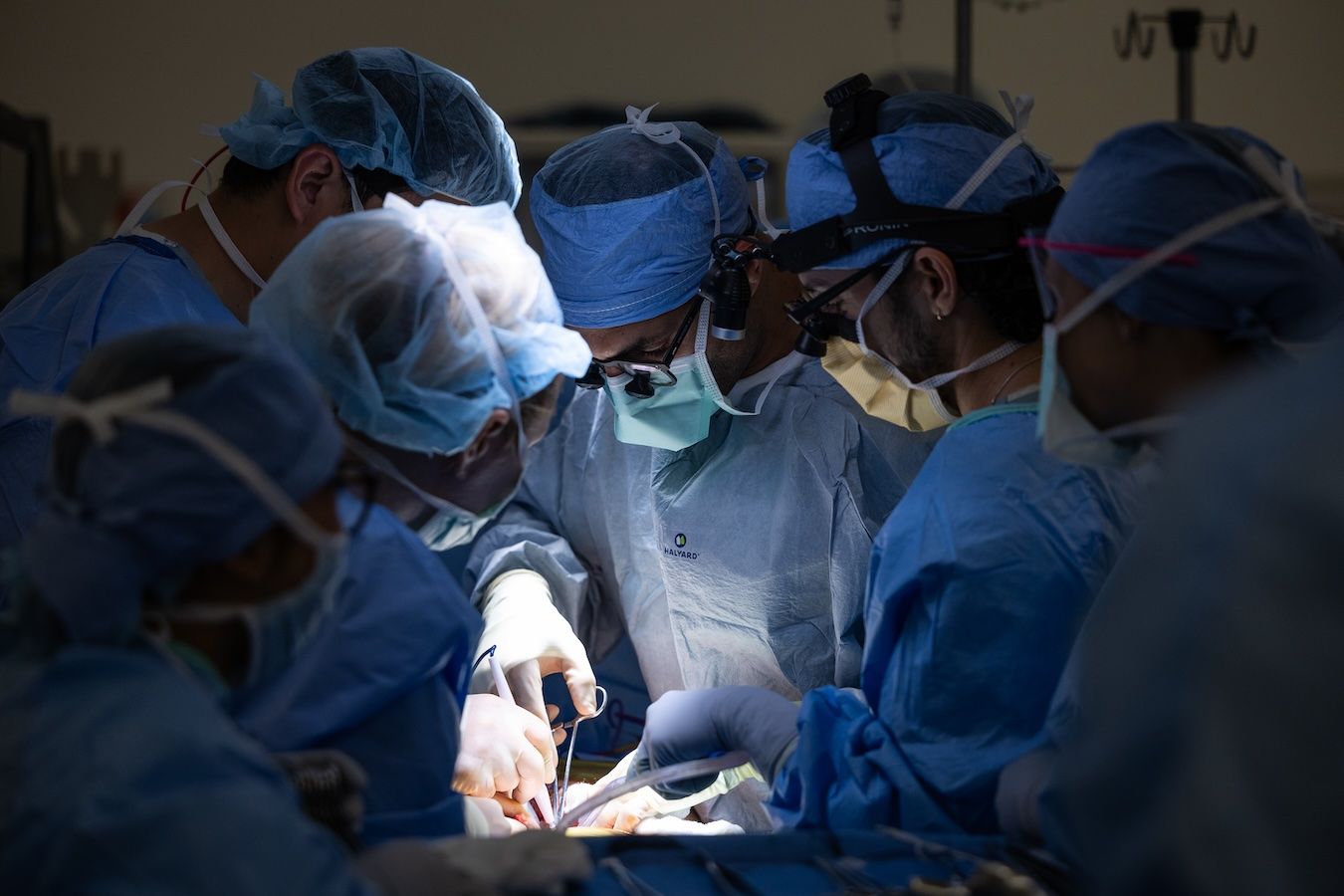 A surgical team in blue gowns, masks, and headlamps performs an operation under bright lights in an operating room.