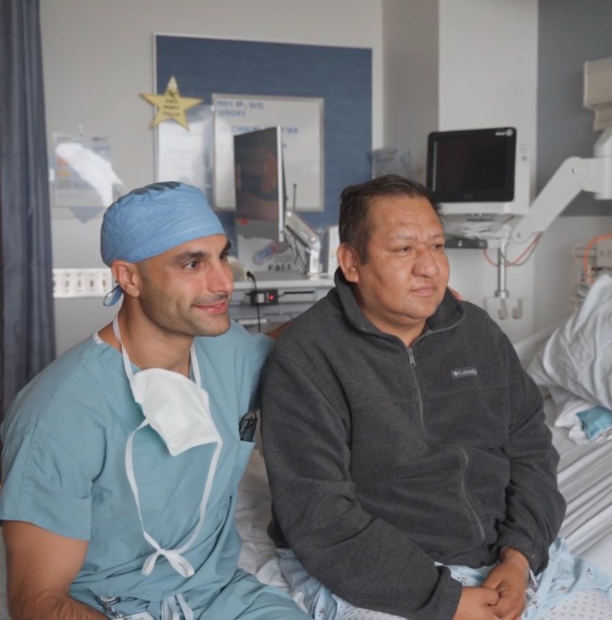 Dr. Nima Nassiri, wearing surgical scrubs and a blue cap, sits beside transplant patient Oscar Larrainzar in a hospital room at UCLA Health. Both men face the camera, with medical equipment visible in the background.