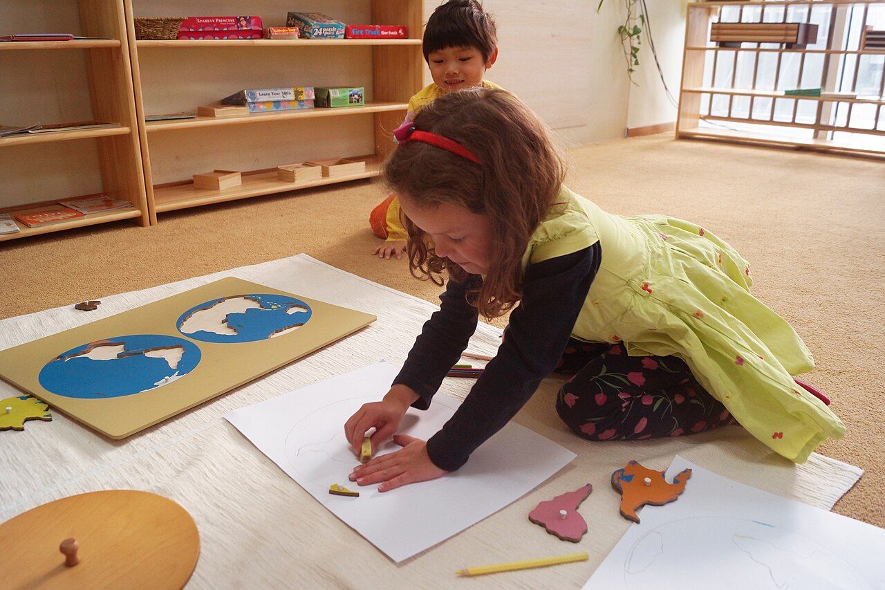 Preschool-aged girl in yellow dress plays on the floor with geography shape puzzle as young boy watches behind her