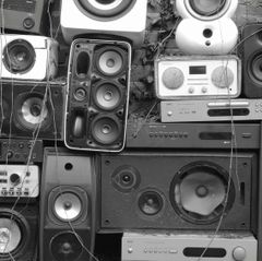 Black and white photo of a wall densely covered with old stereo speakers, boomboxes, and audio equipment of various sizes.