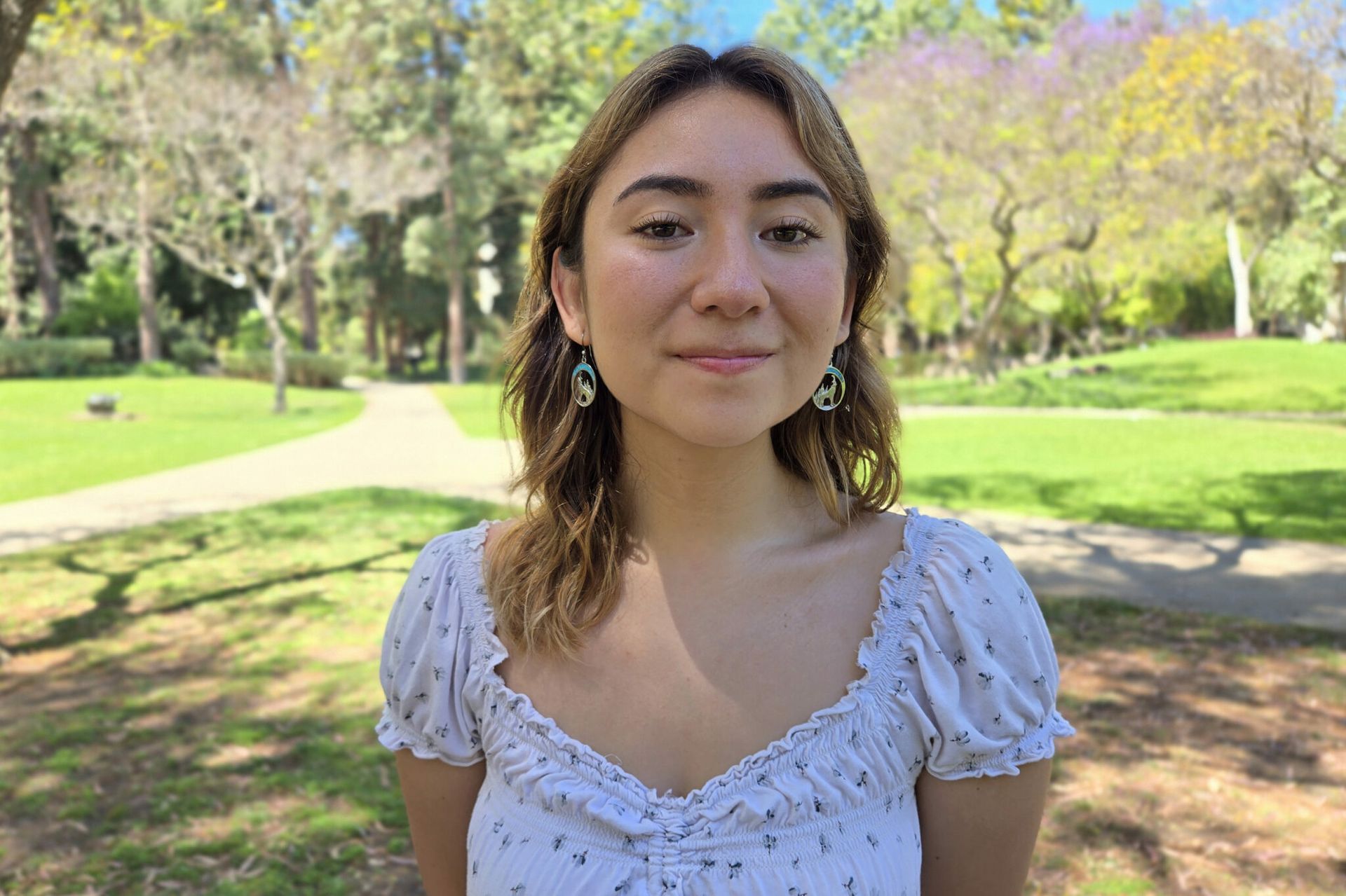 Headshot of Nicolette Bond smiling outdoors on a sunny campus lawn, wearing a white floral top and green earrings.