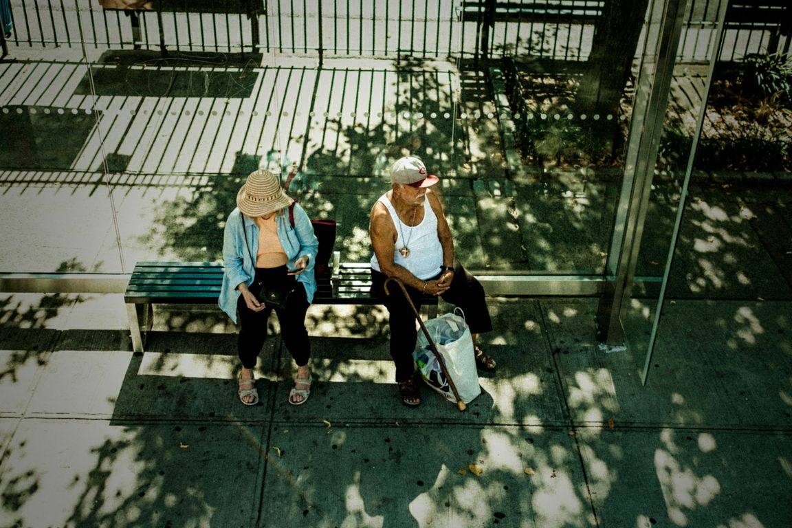 A man and woman sit on the bench of a bus stop covered by tree shade