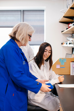 Dr. Samantha Butler stands in a blue lab coat in her laboratory space and shows a blue lab test to a seated Talin Demirjie, who wears a white lab coat.   