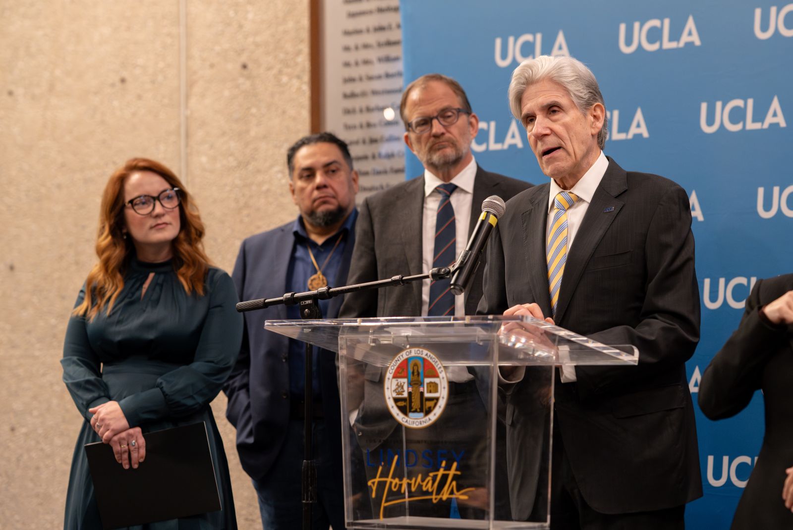 County Supervisor Lindsey Horvath watches as UCLA Chancellor Julio Frenk speaks at a lucite podium in front of blue background with UCLA letters in white