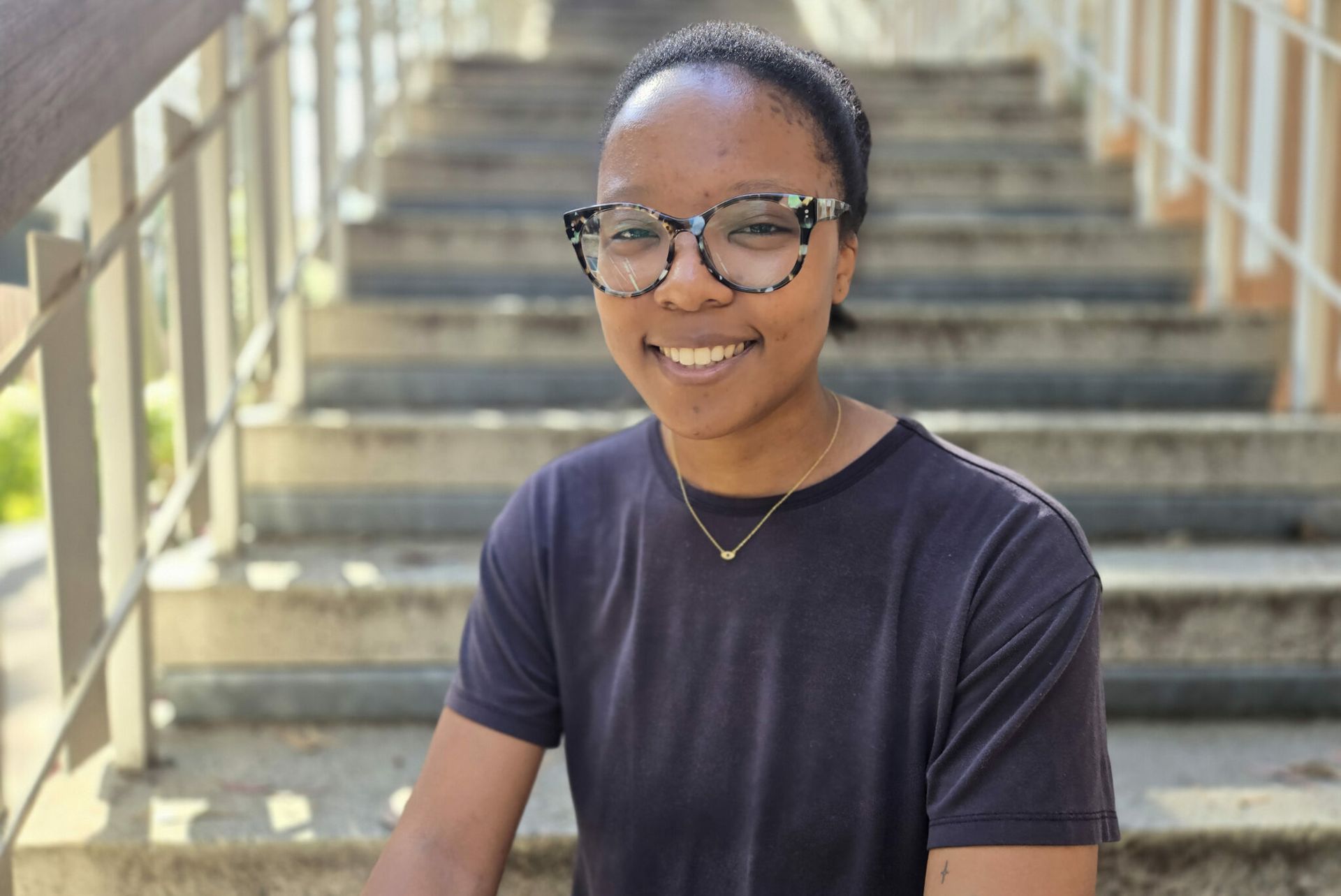 Leila Chiddick sits smiling on outdoor steps in sunlight, wearing a dark blue t-shirt and tortoiseshell glasses.