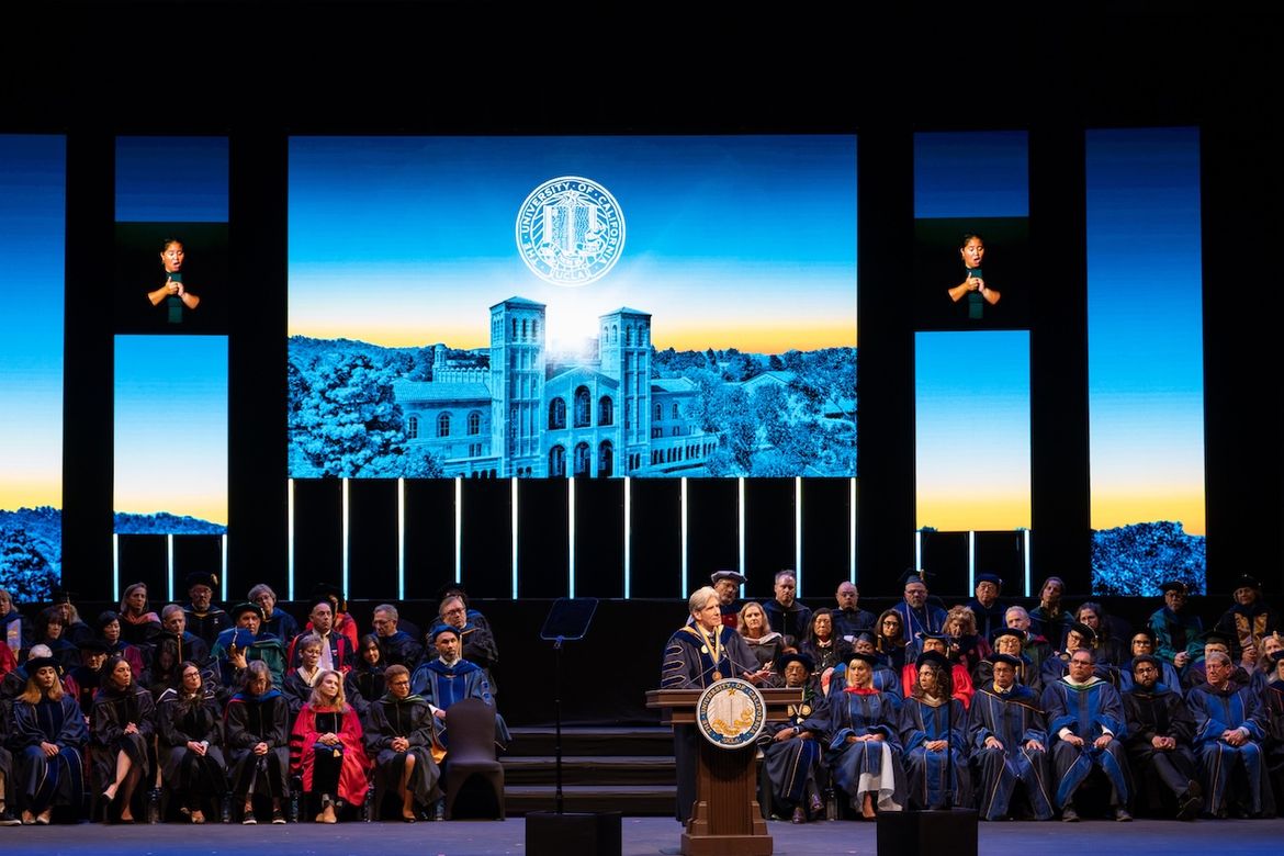 Wide shot of stage as Julio Frenk stands behind a podium. Four rows of guests are seated on each side of the podium.