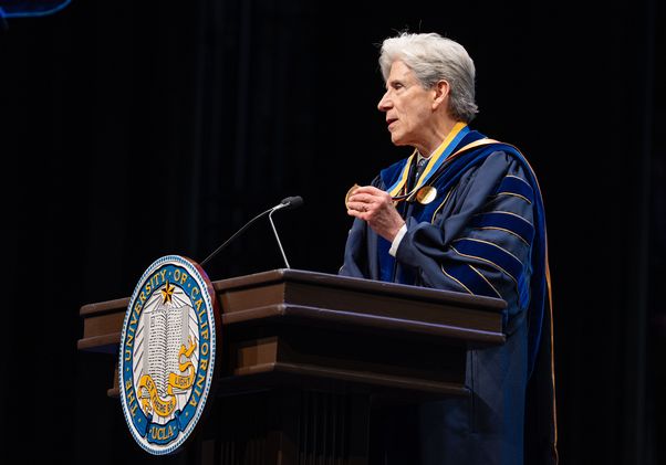 UCLA Chancellor Julio Frenk speaks at a podium wearing full academic regalia, including a blue doctoral gown and medal, during his inauguration ceremony at Royce Hall.