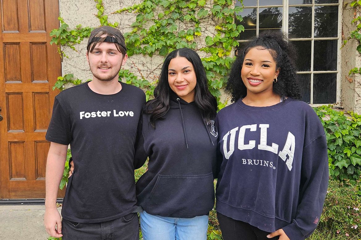 Justin Monk, Anna James, and Soraya Leonard stand together smiling outside an ivy-covered building. Monk wears a "Foster Love" shirt; Leonard wears a UCLA Bruins sweatshirt.