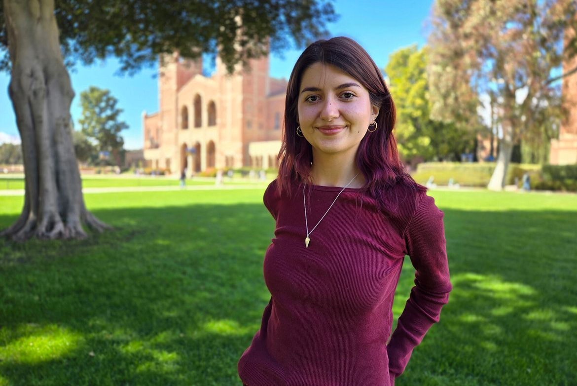 Victoria Gutierrez stands smiling on the UCLA campus lawn with Royce Hall in the background, wearing a burgundy long-sleeve top and a gold necklace.