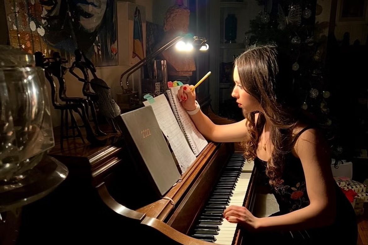 Mia Ruhman sits at an upright piano in a dimly lit room, writing on sheet music by lamplight.