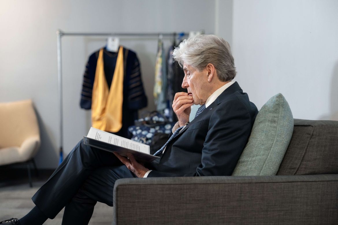 UCLA Chancellor Julio Frenk sits on a sofa reviewing notes in a quiet room, with academic regalia hanging on a rack in the background, as he prepares for his June 5 inauguration.