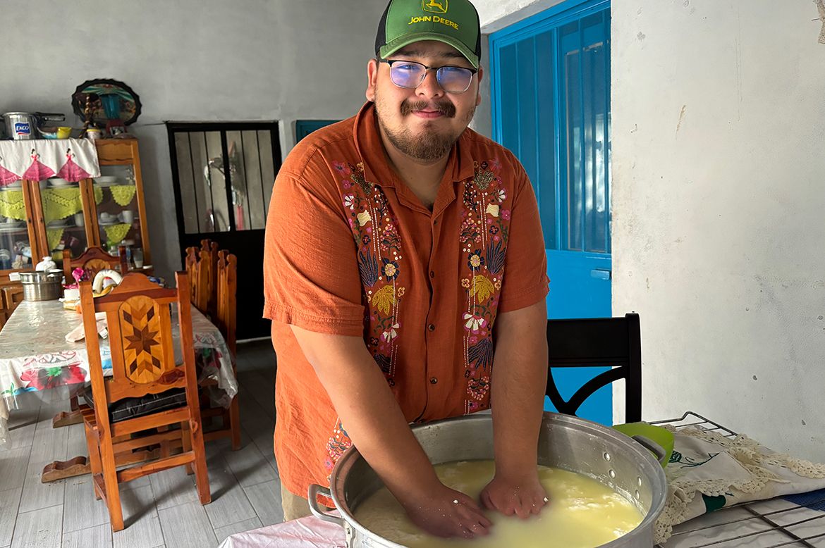 Guillermo Miranda leans over a large metal bowl of dough on a table, smiling, wearing an orange embroidered shirt and a green cap indoors.