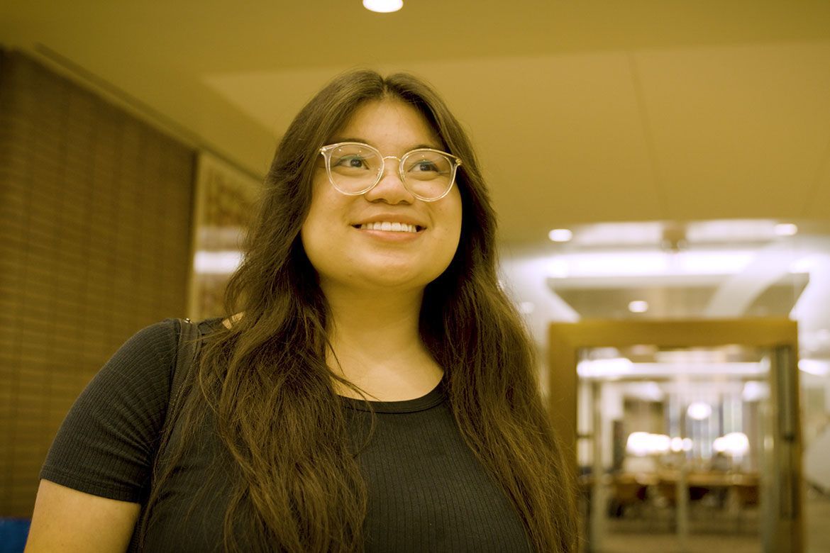 Headshot of Evelyn Giron smiling in a warmly lit indoor hallway, wearing glasses and a black top.