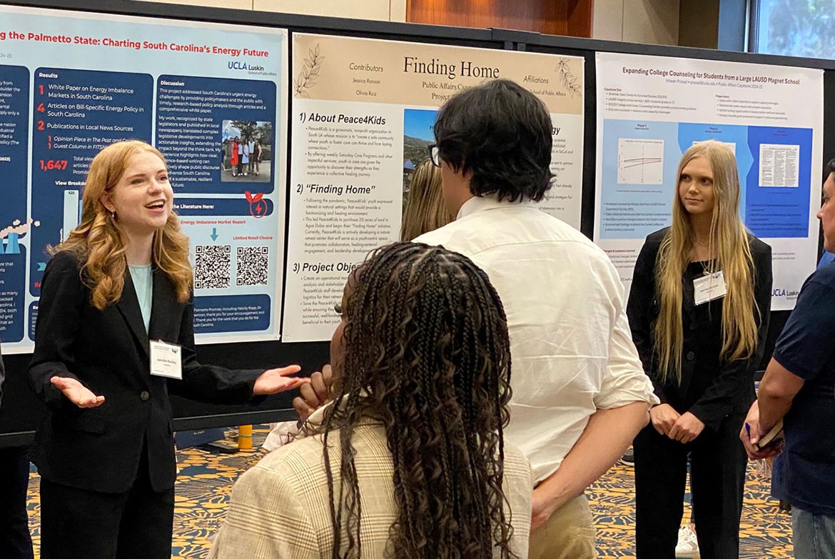 Jennifer Buckley, far left, presents her research poster to a small group of attendees at an academic conference, gesturing animatedly beside displayed project boards.
