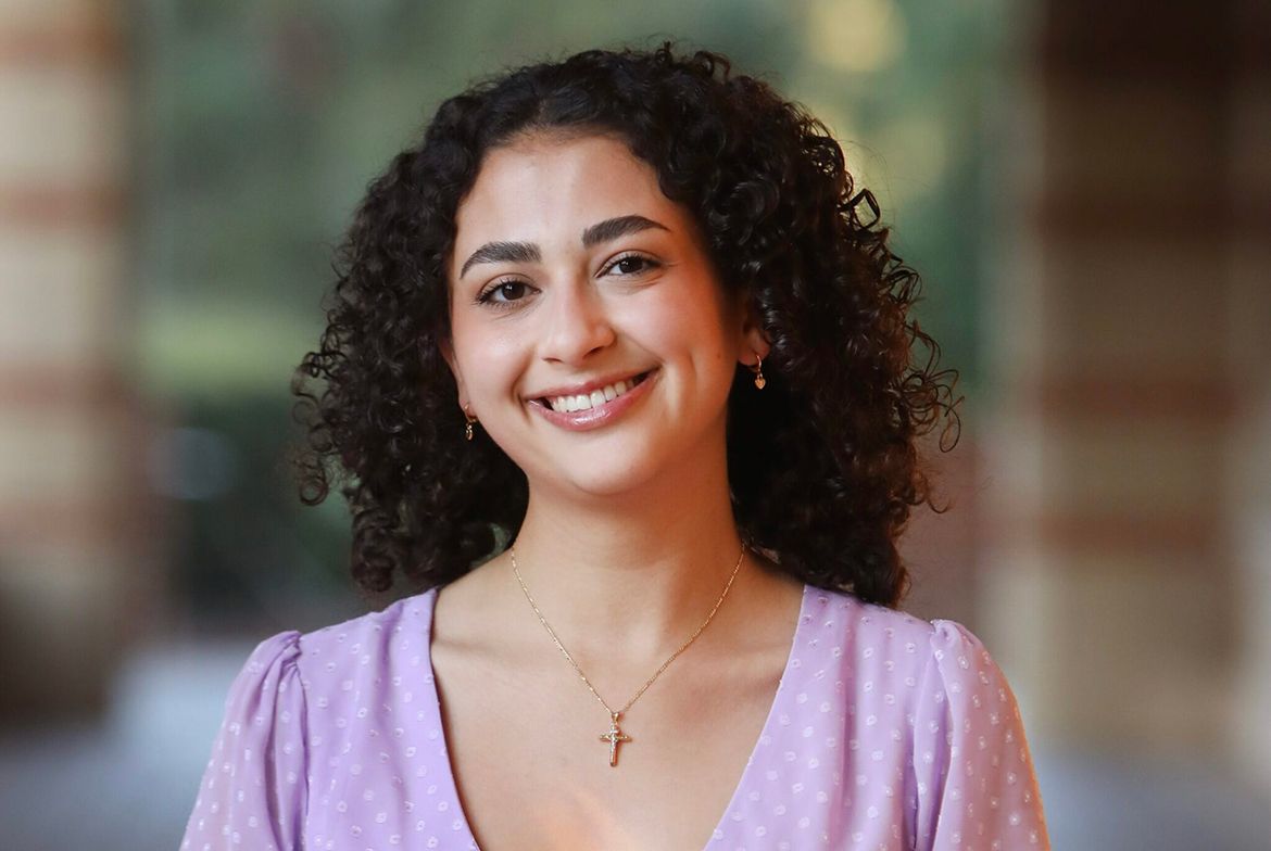 Headshot of Katie Cunningham smiling, wearing a lavender top and a gold cross necklace, photographed against a warm brick background.