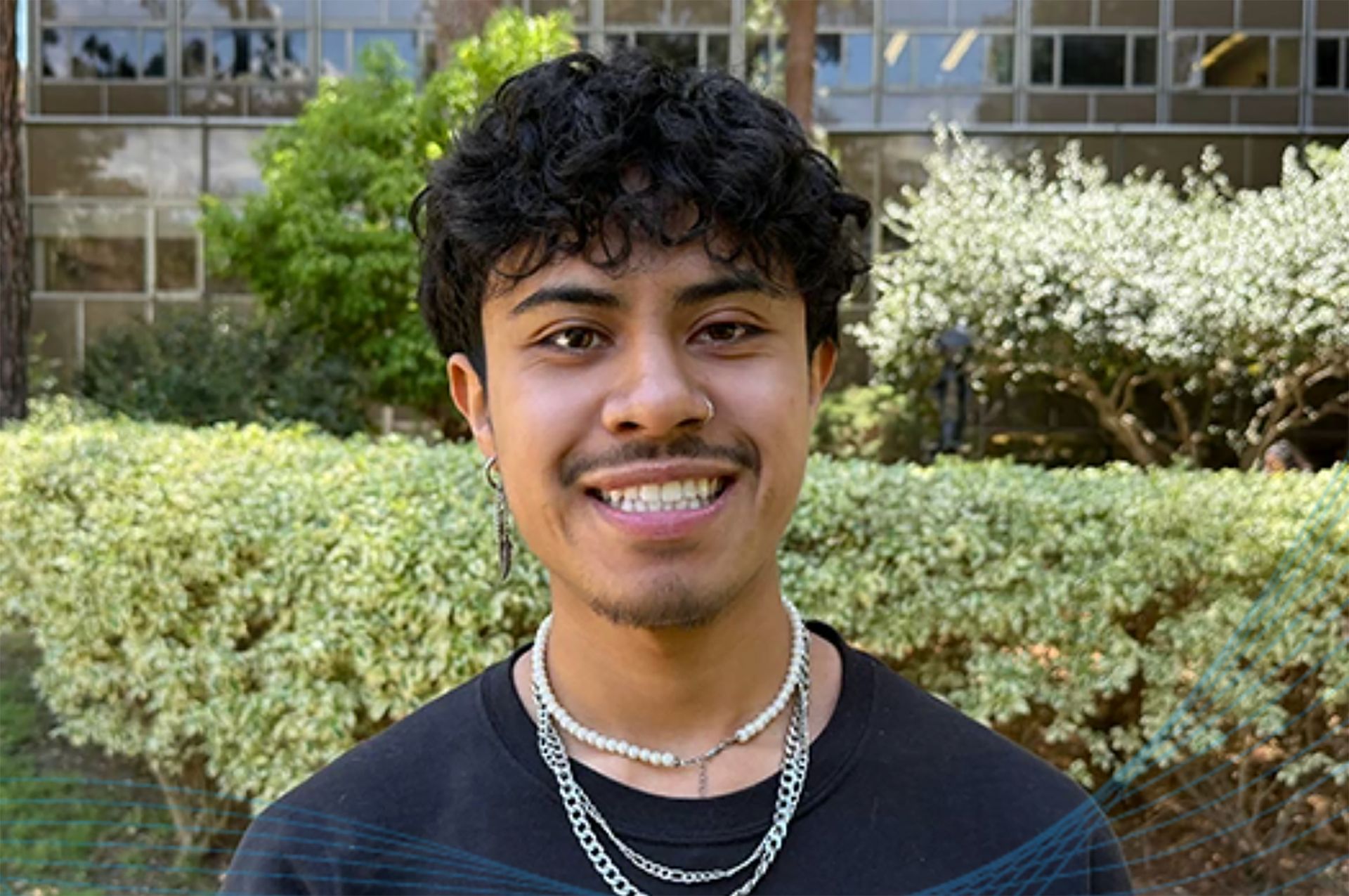 Headshot of Joaquin Garcia smiling outdoors on the UCLA campus, wearing a dark shirt and layered silver necklaces, with green shrubs in the background.